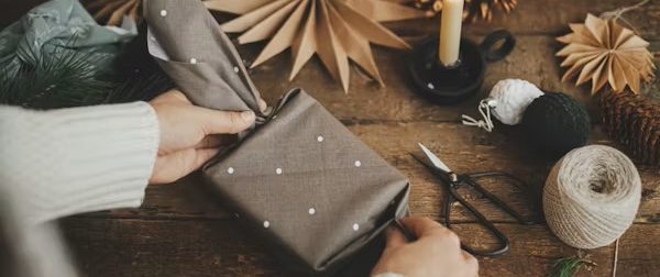 Person wrapping a gift in brown material with a nature theme Person wrapping a gift in brown material with a nature theme in the background, with scissors, string and Christmas decorations around the g